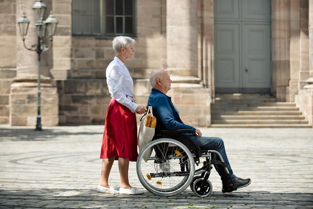 Eine Seniorin schiebt ihren Mann, der im Rollstuhl sitzt, über einen Platz