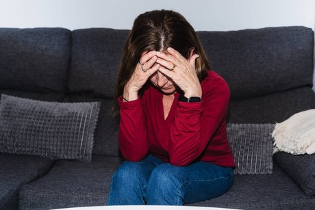 Eine ältere Frau sitzt auf einem Sofa, hat den Kopf gesenkt und hält sich die Hände vor das Gesicht. Sie sieht erschöpft oder traurig aus. 