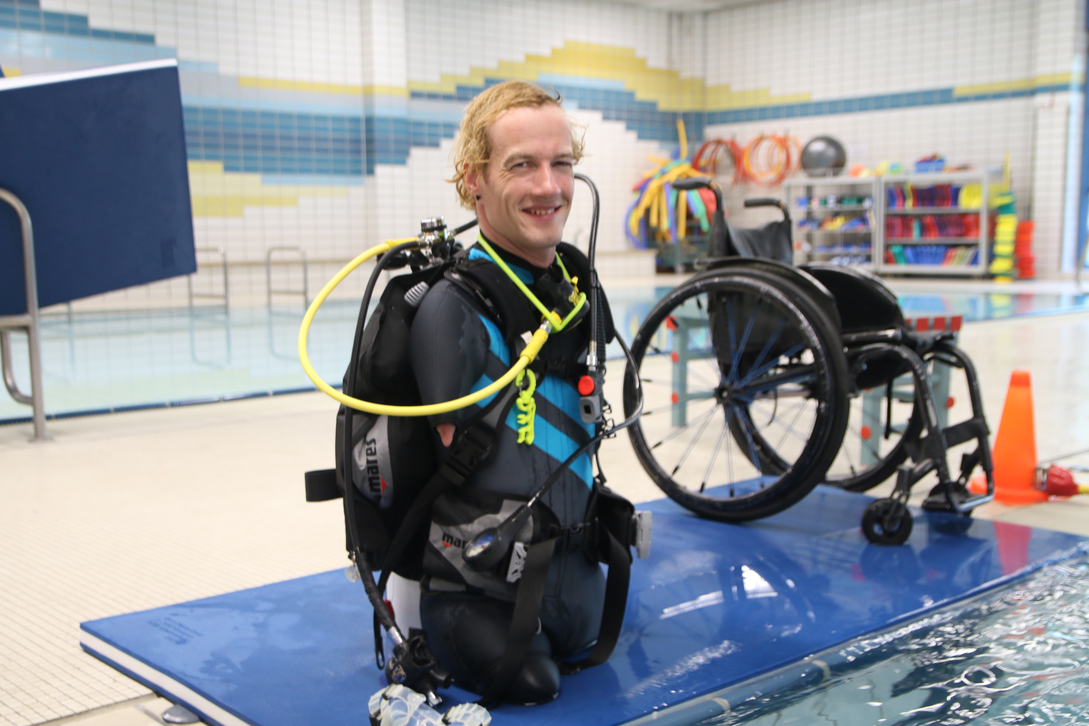 Janis McDavid am Beckenrand im Schwimmbad, er kniet auf einer blauen Matte. Im Hintergrund steht ein Rollstuhl. 