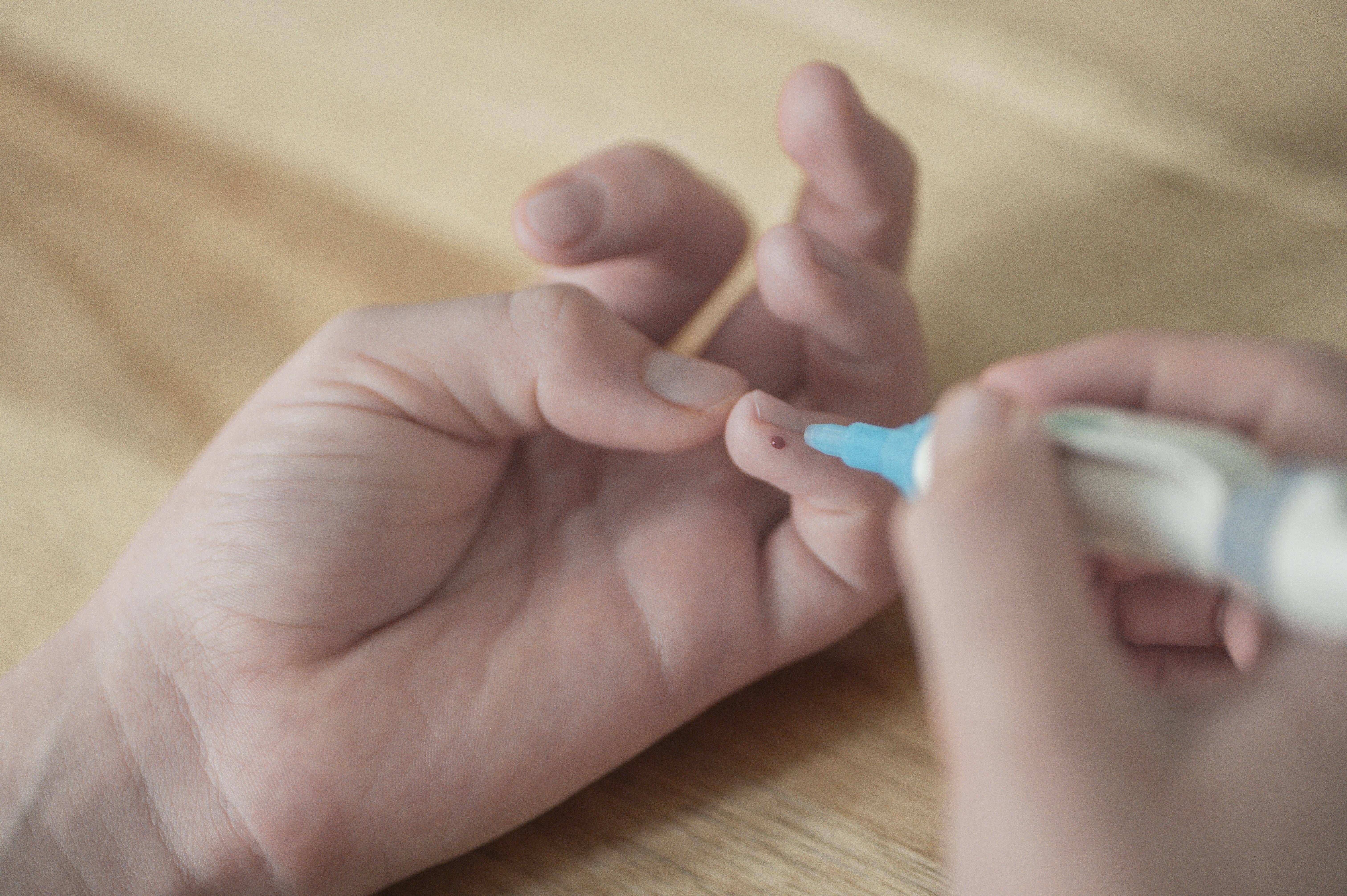 Das Bild zeigt eine Kinderhand, deren kleiner Finger einen Tropfen Blut zeigt. In der rechten Hand hält das Kind die Lanzette als Stechhilfe, hat sich gerade gepiekst, um den Blutzucker zu bestimmen.