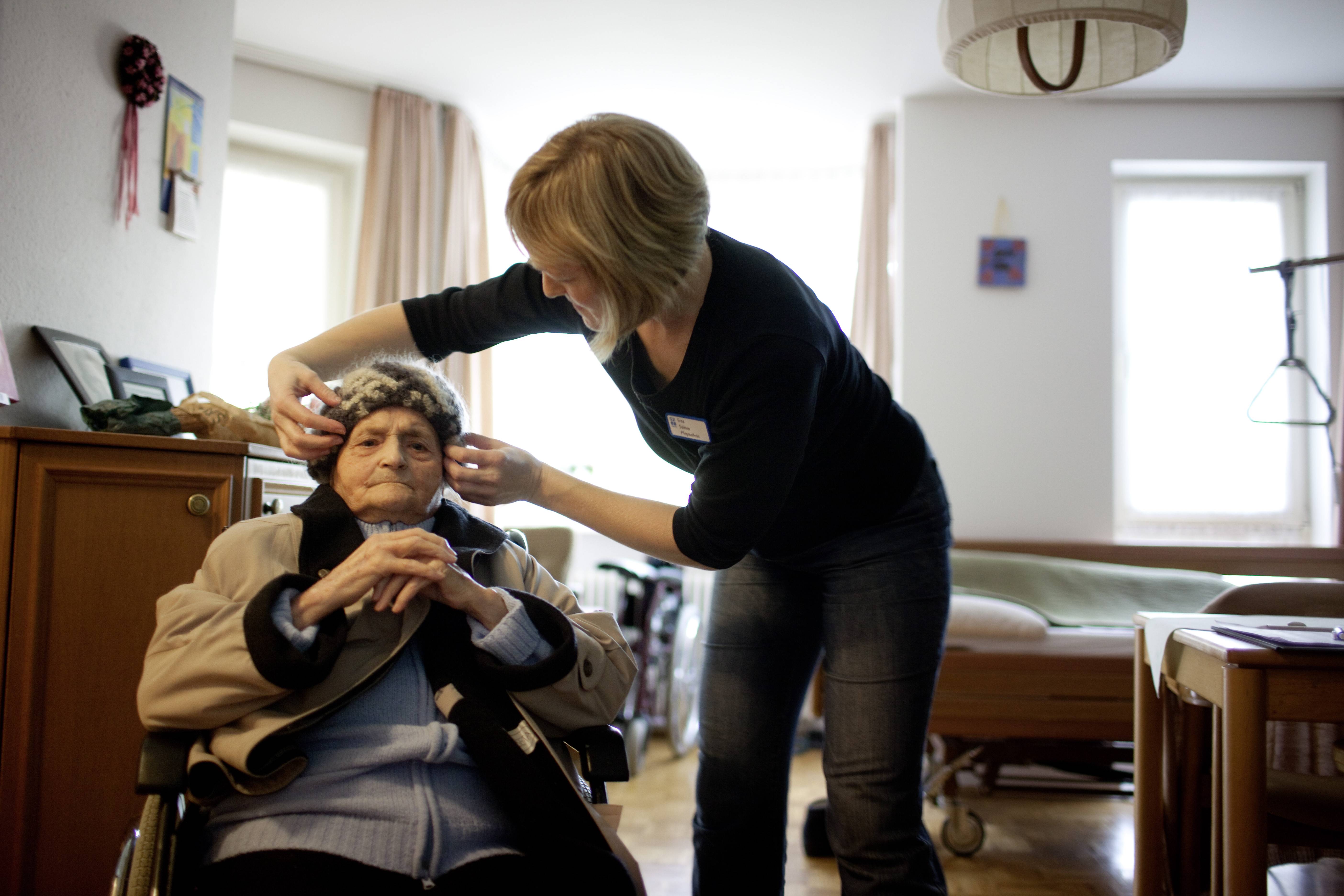 Eine alte Dame im Rollstuhl in ihrem Pflegeheim-Zimmer, eine jüngere Altenpflegerin hilft ihr gerade, ihre Mütze aufzusetzen.