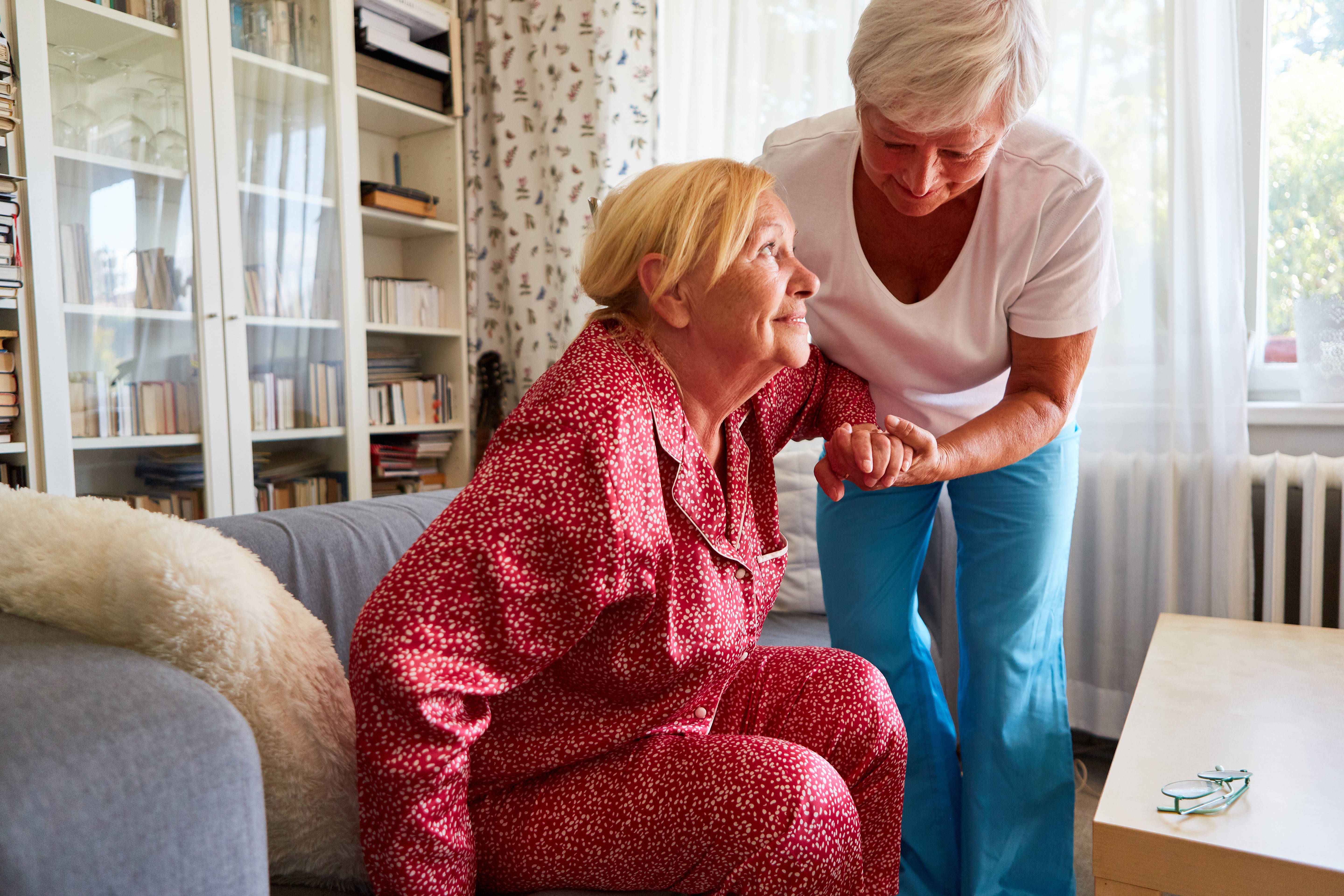 Symbolfoto für Pflege zu Hause: Eine ältere Frau hilft einer anderen Frau, die einen Pyjama trägt, beim Aufstehen von einem Sofa.