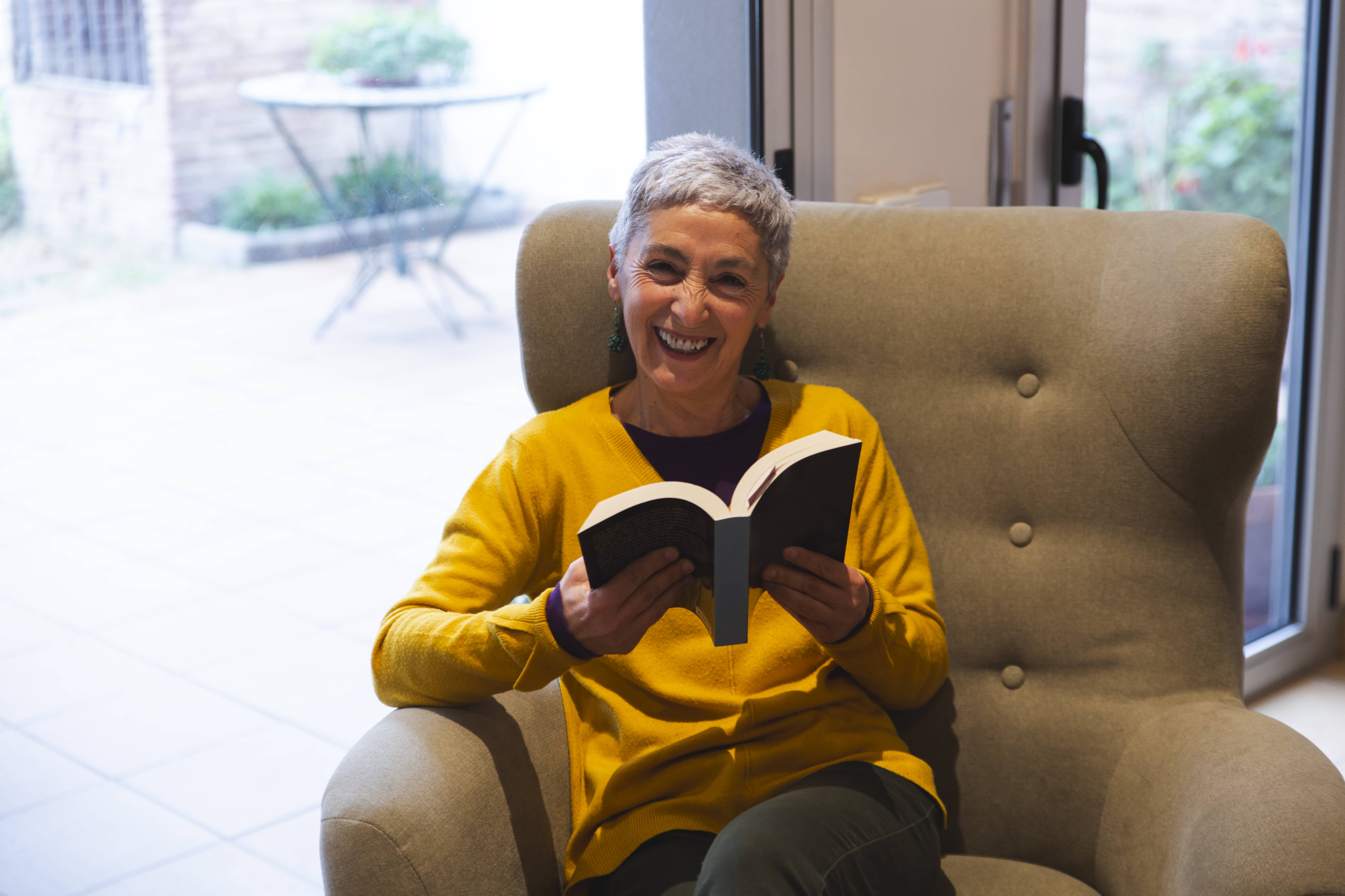 Eine ältere Frau sitzt in einem Sessel, sie hält ein aufgeschlagenes Buch in den Händen, lacht in die Kamera